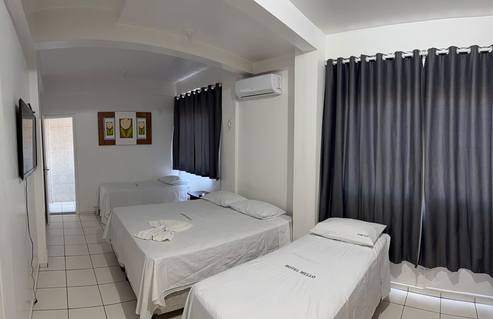 Quarto do Hotel Mello com TV na parede e banheiro privativo ao fundo